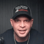 Man wearing black baseball cap with white text speaking to camera in front of dark wall