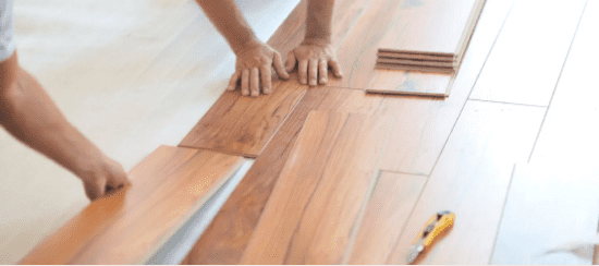 Hands installing wooden laminate flooring planks on white subfloor surface