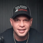 Man wearing black baseball cap with white text speaking to camera in front of dark wall