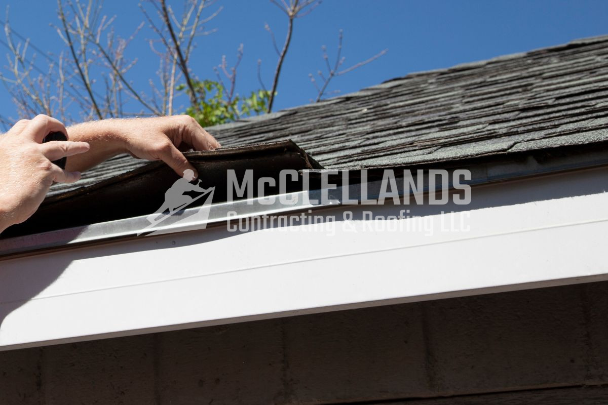 Hands repairing shingles on a residential roof under clear blue sky.