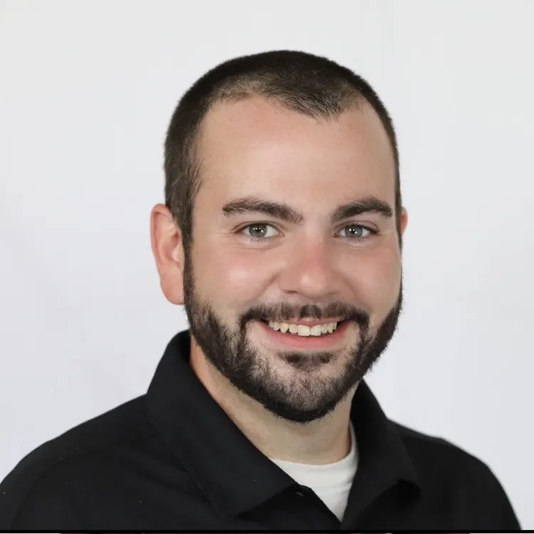 Smiling man with beard and dark shirt against plain background.